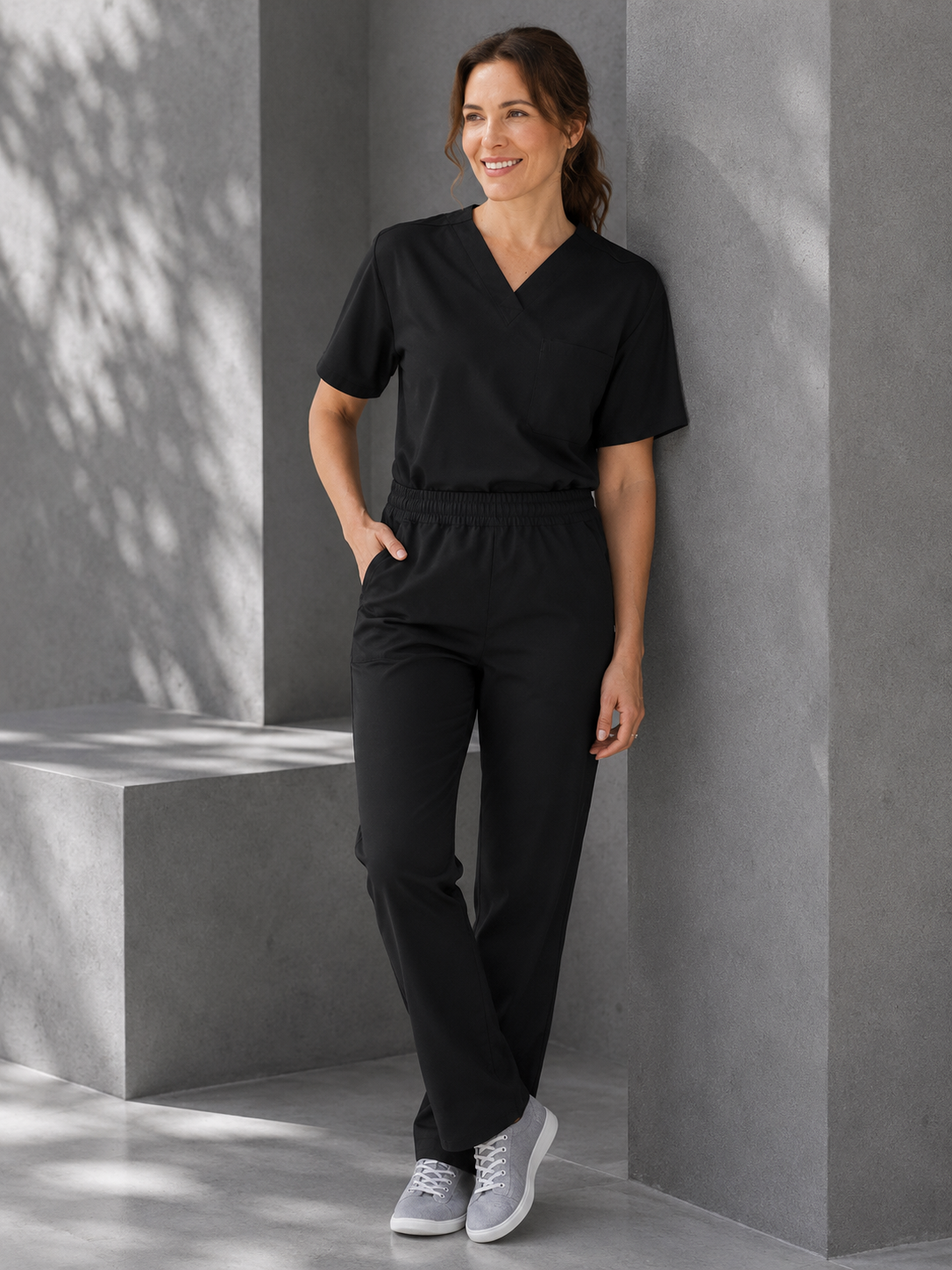 A woman wearing a black V-neck scrub top and matching black scrub pants, standing against a gray concrete wall in a modern architectural setting.