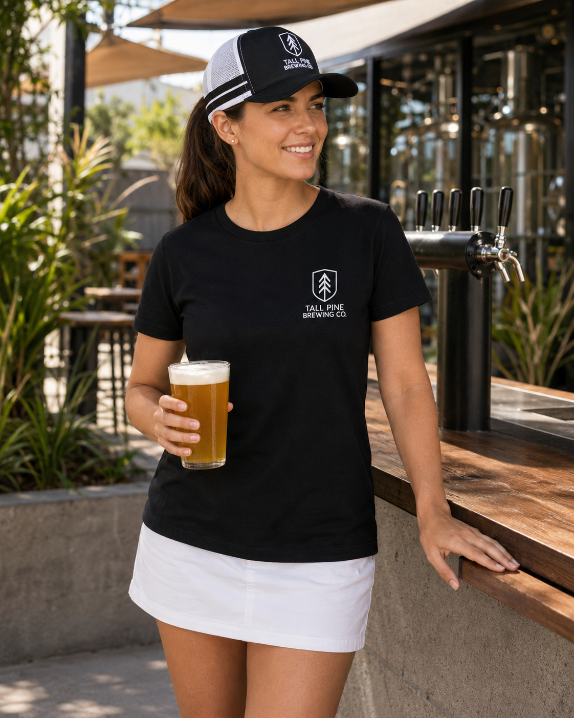 A woman stands at an outdoor bar holding a glass of beer, wearing a black short-sleeve T-shirt with a small white chest logo that reads 'TAX FREE BREWING CO.' and a black-and-white baseball cap.