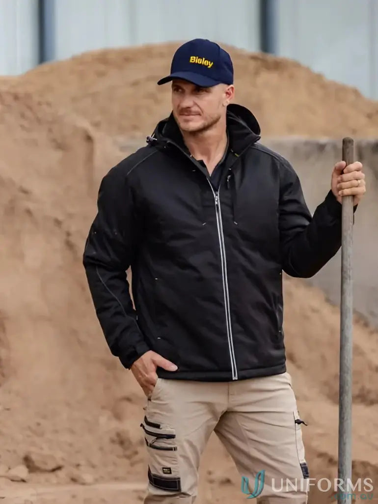 Man in black heated jacket with hood, tan work pants, and blue cap for weather protection
