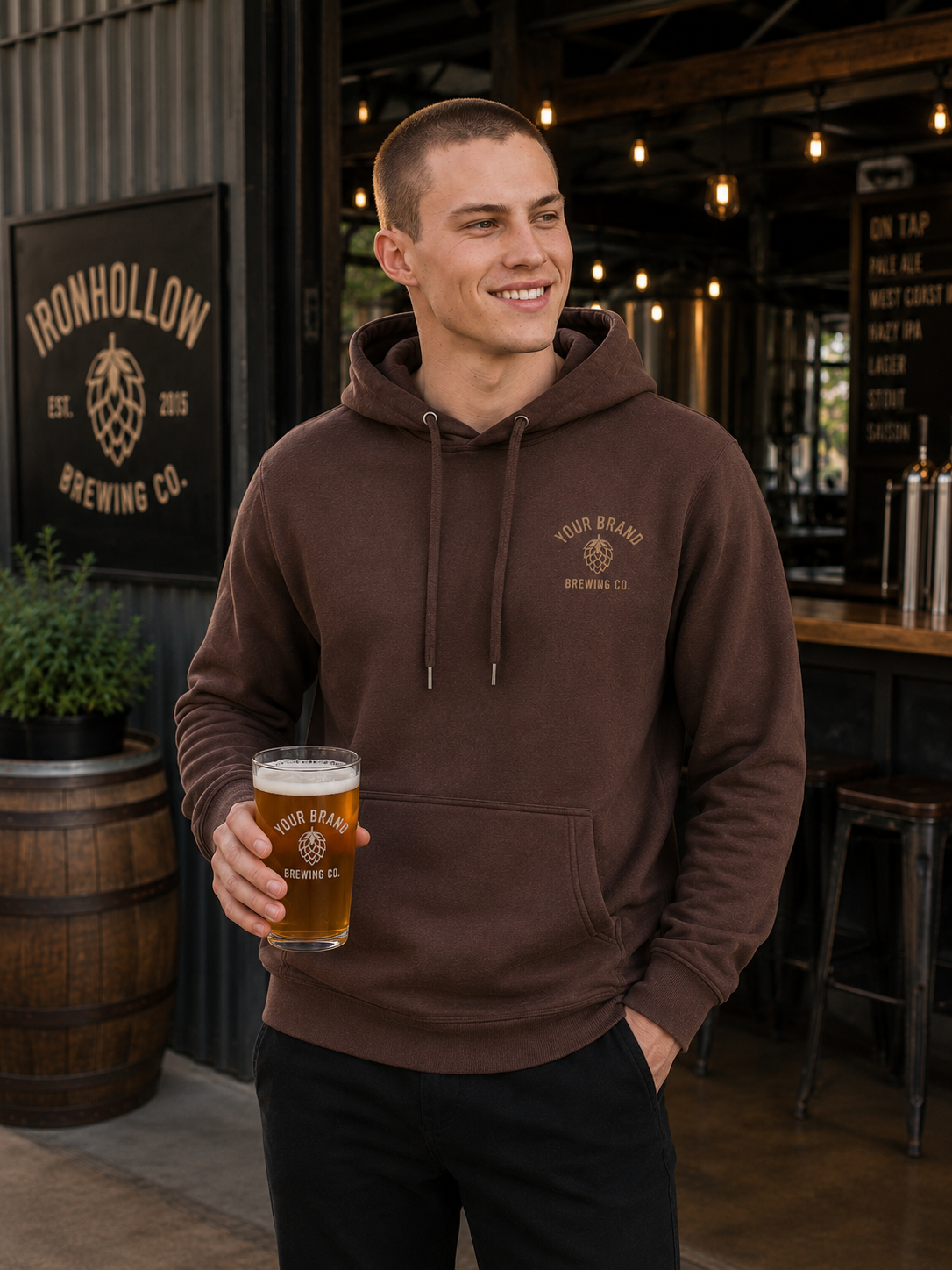 A man wearing a brown pullover hoodie with a drawstring hood and kangaroo pocket, holding a glass of beer in a brewery setting. The hoodie features a gold embroidered logo on the left chest that reads 'YOUR BRAND BREWING CO.