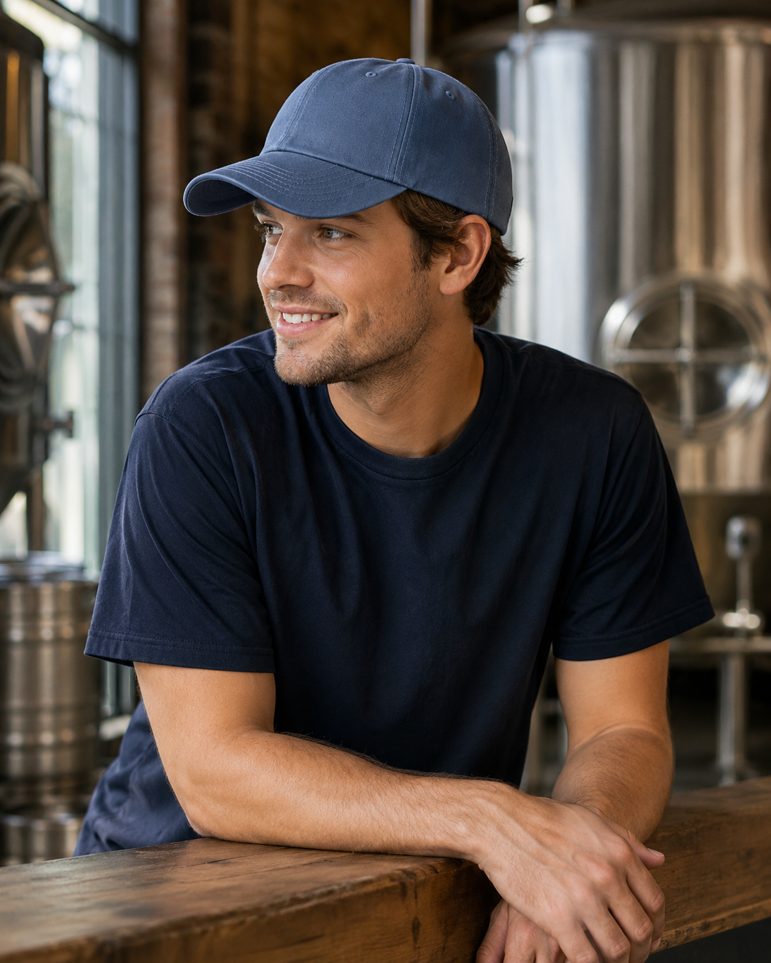 A man wearing a navy blue crewneck t-shirt and a matching navy blue baseball cap, leaning on a wooden counter in a brewery setting with stainless steel fermentation tanks in the background.