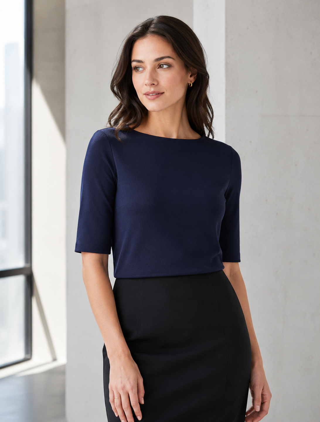 A woman wearing a navy blue short-sleeve knit top and a black pencil skirt, standing in a bright, modern room with a concrete wall and window.