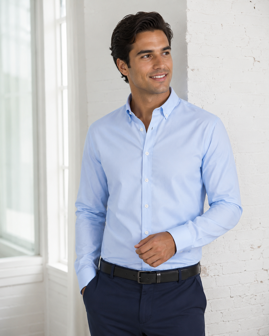A man wearing a light blue long-sleeve button-down dress shirt, navy trousers, and a black belt, standing against a white brick wall.