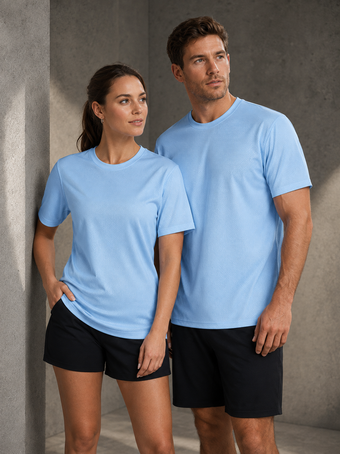 A man and a woman modeling matching light blue crewneck t-shirts and black athletic shorts, standing against a textured gray concrete wall.
