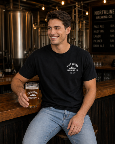 A smiling man in a bar setting wearing a black crewneck t-shirt with a white circular logo that reads 'YOUR BRAND BREWING CO. EST.