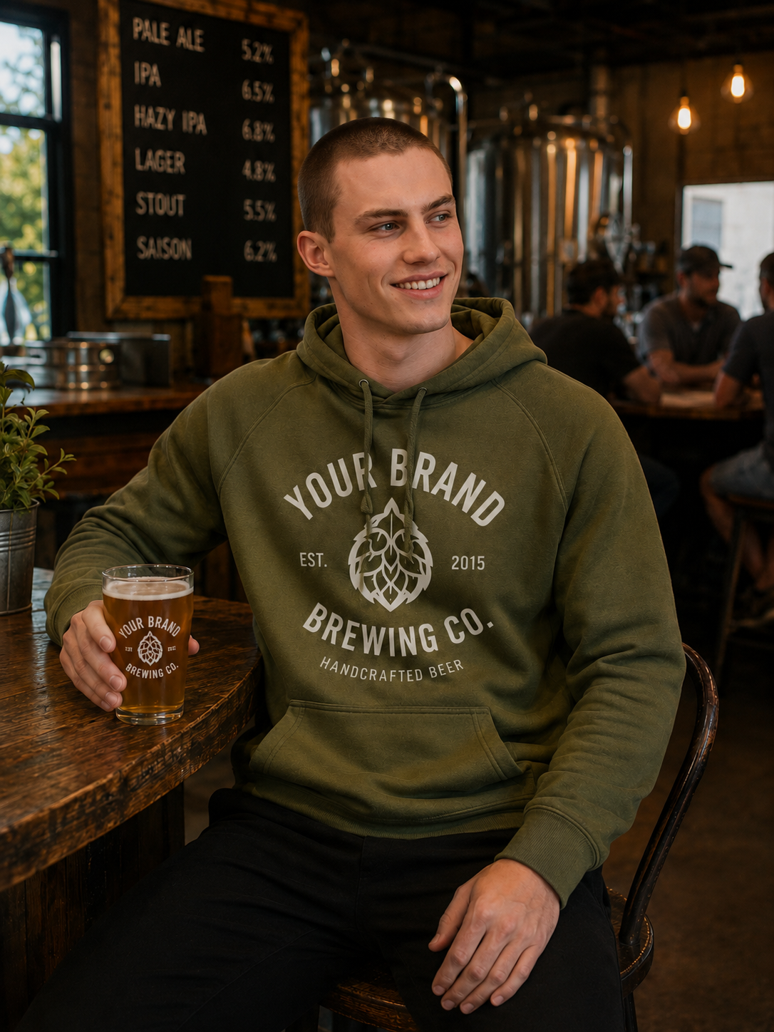 A man sitting in a brewery holding a glass of beer, wearing an olive green hoodie with a white graphic that reads 'YOUR BRAND BREWING CO. HANDCRAFTED BEER'.