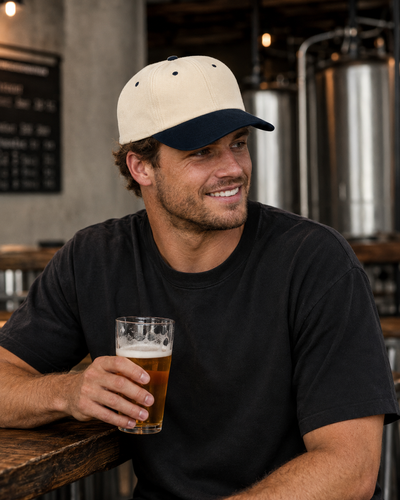 A man in a black t-shirt and a cream and navy baseball cap holding a glass of beer, sitting in a brewery setting with stainless steel fermentation tanks in the background.