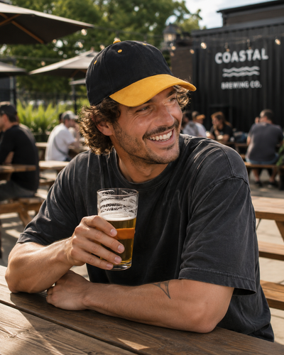 A smiling man at an outdoor bar or brewery seating area holds a pint of beer in a clear glass. He wears a black and yellow baseball cap and a dark gray t-shirt.