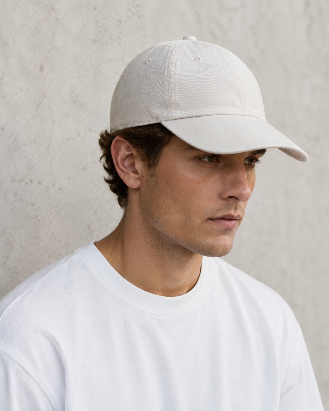 A man wearing a light beige baseball cap and a white t-shirt, photographed from the chest up against a textured light gray concrete wall.