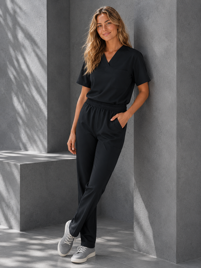A woman wearing a black V-neck scrub top and matching black scrub pants, paired with gray sneakers, posing against a modern gray concrete wall with soft shadows.