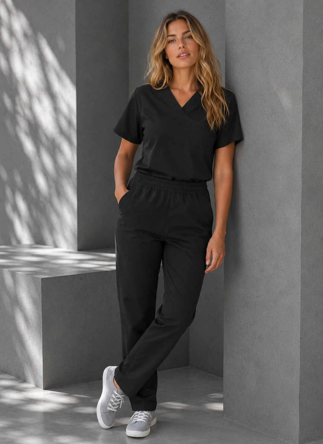 A woman wearing a black V-neck short-sleeve scrub top and matching black scrub pants, paired with white sneakers, posing in a modern gray concrete setting with dappled sunlight.