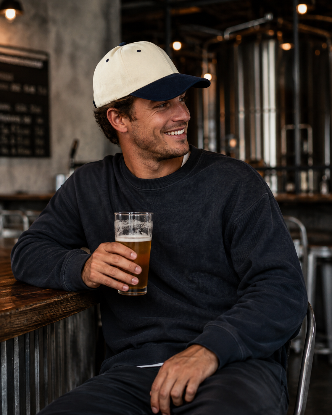 A man sitting in a brewery or bar, wearing a navy blue crewneck sweatshirt and a cream and navy baseball cap, holding a glass of beer.