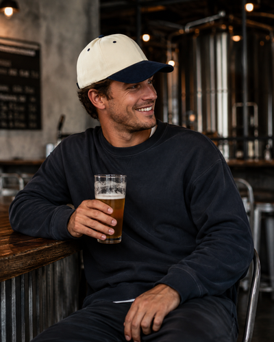A man sitting in a brewery or bar, wearing a navy blue crewneck sweatshirt and a cream and navy baseball cap, holding a glass of beer.