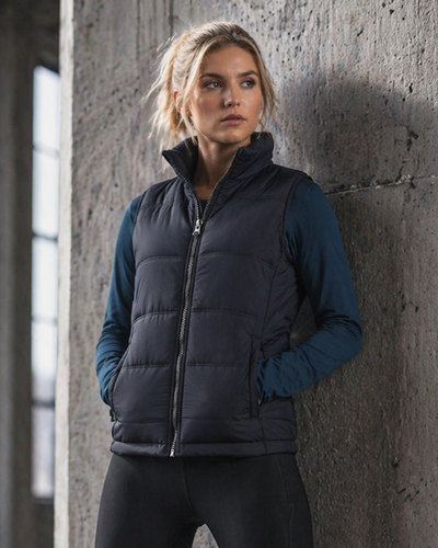 A woman wearing a dark navy quilted puffer vest over a dark blue long-sleeve shirt and black leggings, leaning against a textured concrete wall.