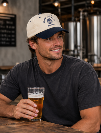 A smiling man in a dark gray t-shirt sits at a wooden table in a brewery, holding a glass of beer. He is wearing a cream and navy blue baseball cap with the text 'Coastal Brewing Co EST.