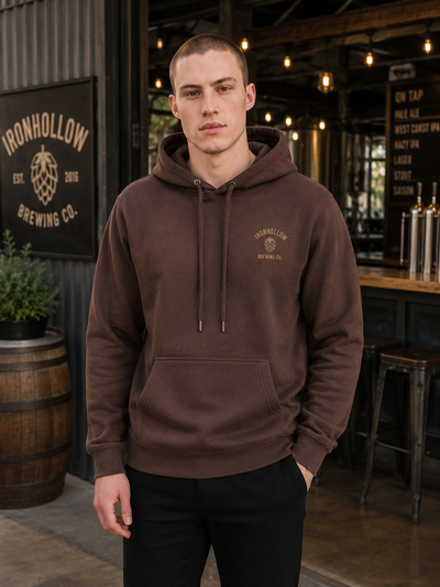 A man wearing a brown pullover hoodie with a drawstring hood and a kangaroo pocket, standing in a brewery setting. The hoodie features a small embroidered 'Ironhollow Brewing Co.