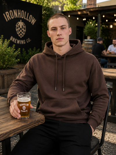 A man sitting at an outdoor patio table wearing a brown pullover hoodie with drawstrings and a kangaroo pocket, holding a glass of beer. A black sign in the background reads 'IRONHOLLOW BREWING CO.