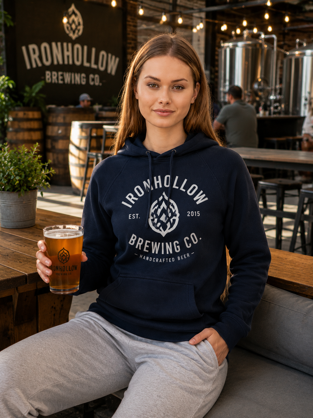 A woman sits at a outdoor brewery patio holding a glass of beer, wearing a navy blue hoodie with a drawstring hood and kangaroo pocket, paired with light gray sweatpants. The background features wooden tables, other patrons, and a black sign reading 'IRONHOLLOW BREWING CO.