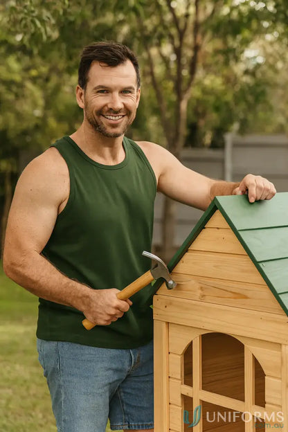 Smiling man in green workwear tank top holding hammer next to wooden playhouse with green roof