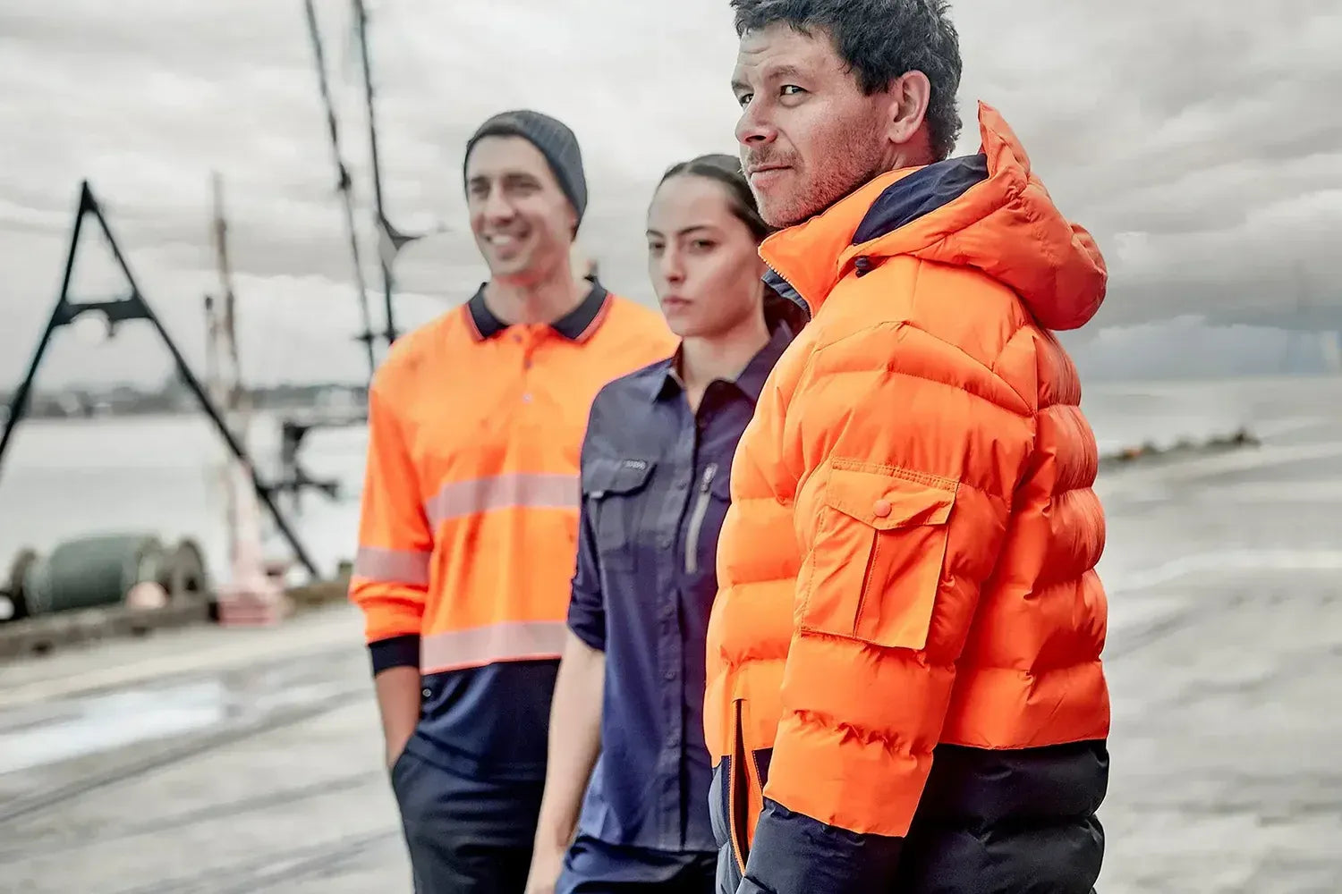 Three people wearing orange safety jackets standing outdoors with a cloudy sky and industrial background.