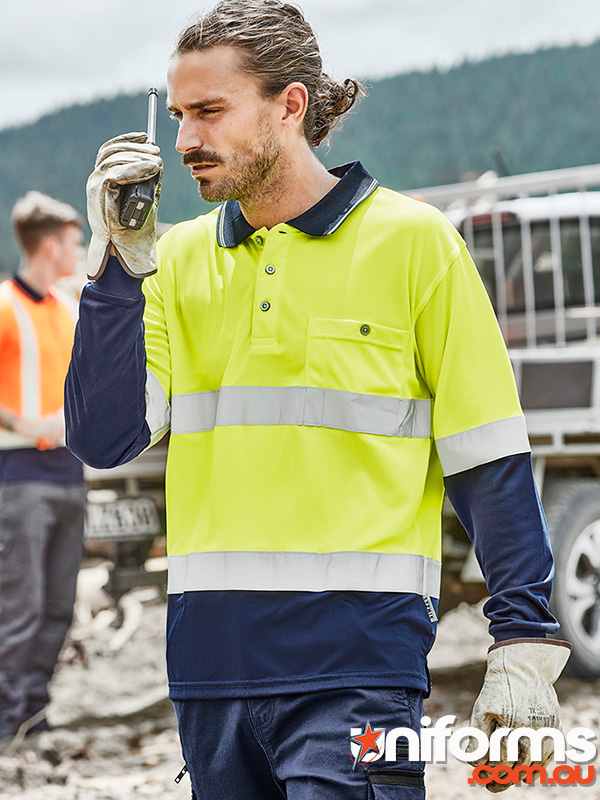 A man wearing a  yellow and blue Syzmic Hi Vis Polo, converses on a cell phone outdoors.