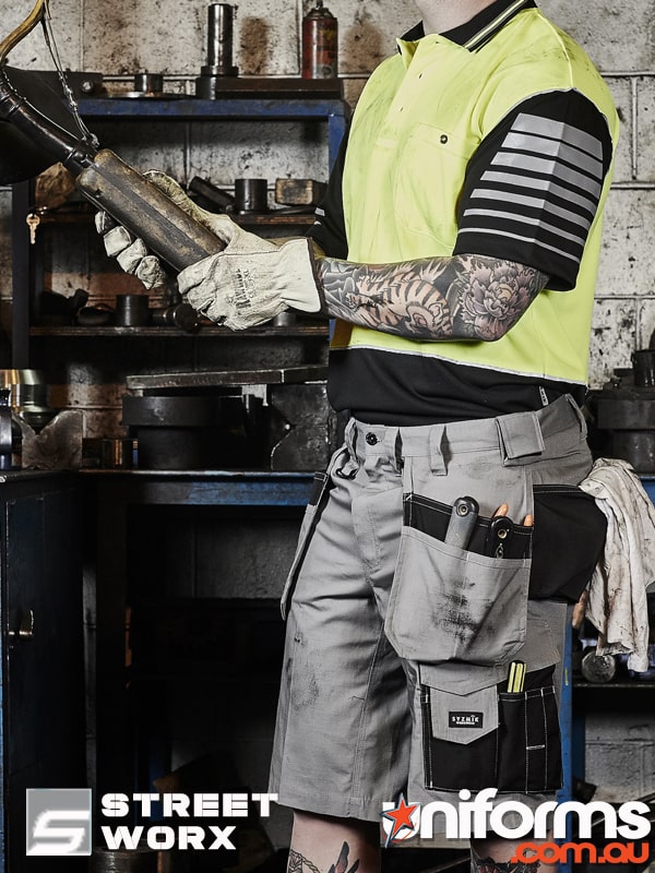A man in a yellow polo and Syzmic multi-pocket shorts, style ZS510 holding a tool in a workshop..