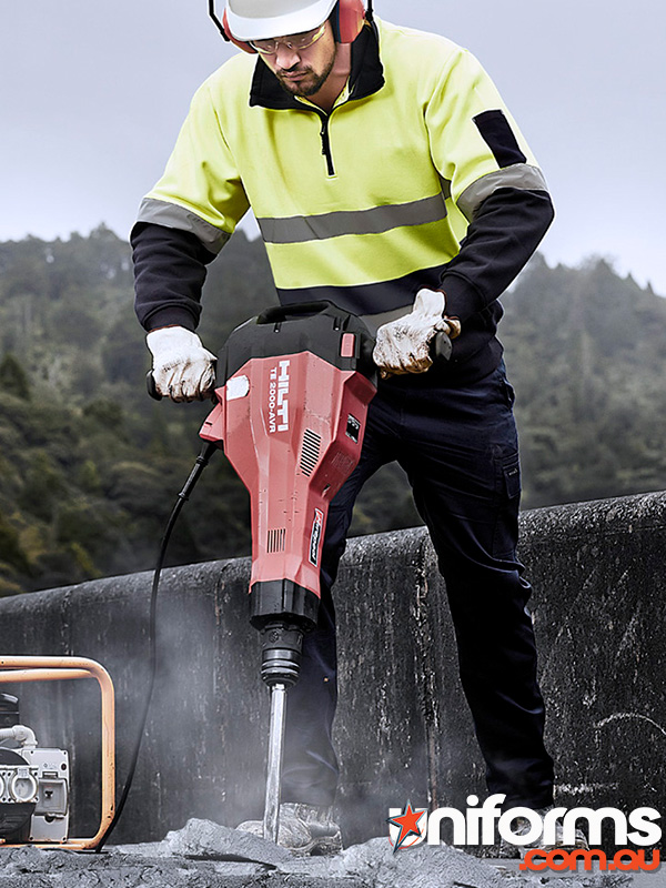 A man wearing a yellow Syzmic 1/4 zip pullover is using a hammer drill to break concrete,demonstrating construction work.